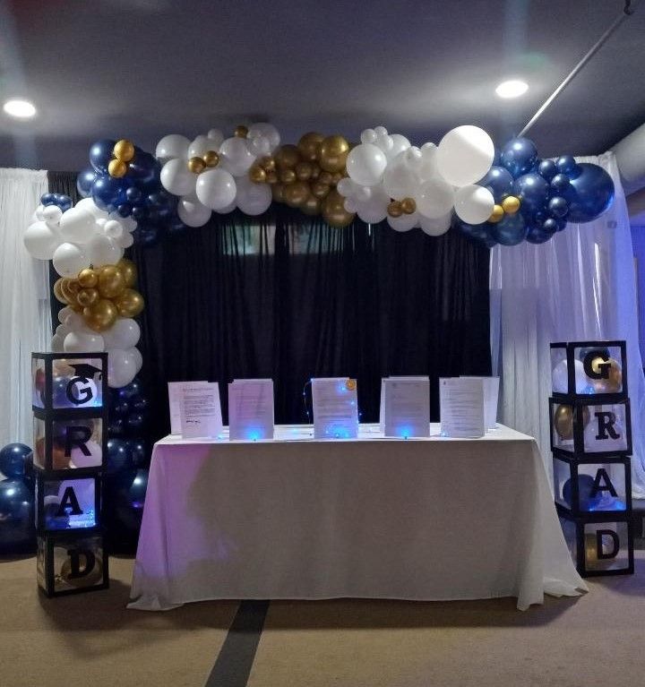 Graduation party setup with a balloon arch in blue, white, and gold, and a table with glowing displays.