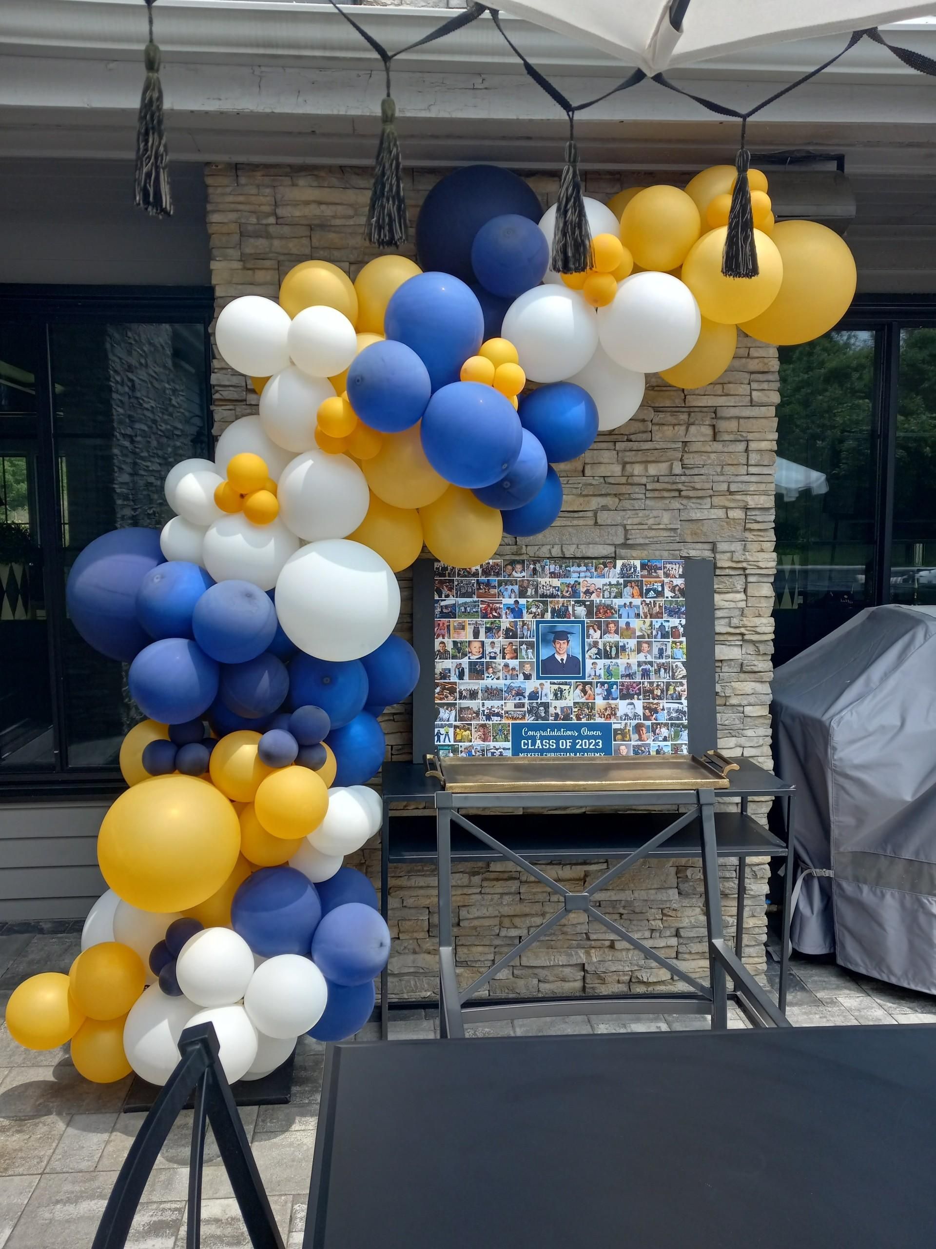 Graduation celebration: Balloon arch in blue, white, and gold; photo display on table.