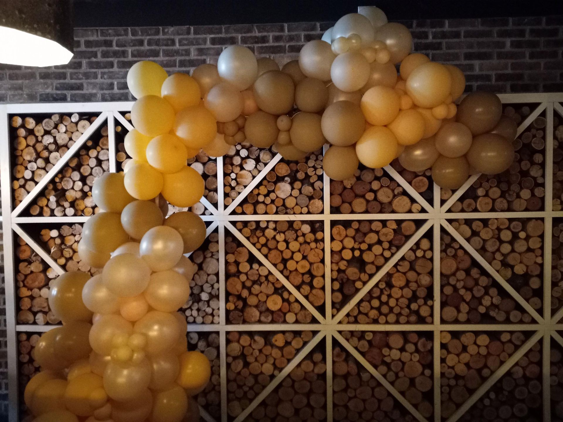 Yellow and brown balloon arch over a white lattice, in front of a wood-pile wall.