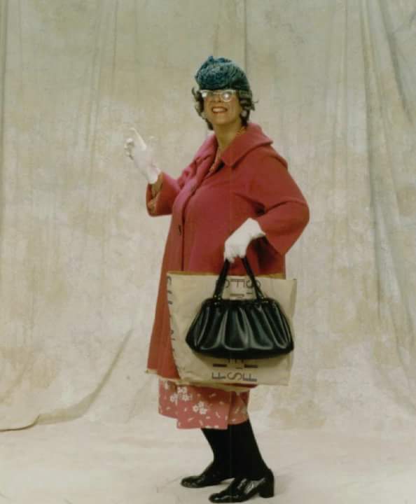 Woman in pink coat, hat, and gloves holding a bag, standing against a neutral backdrop.