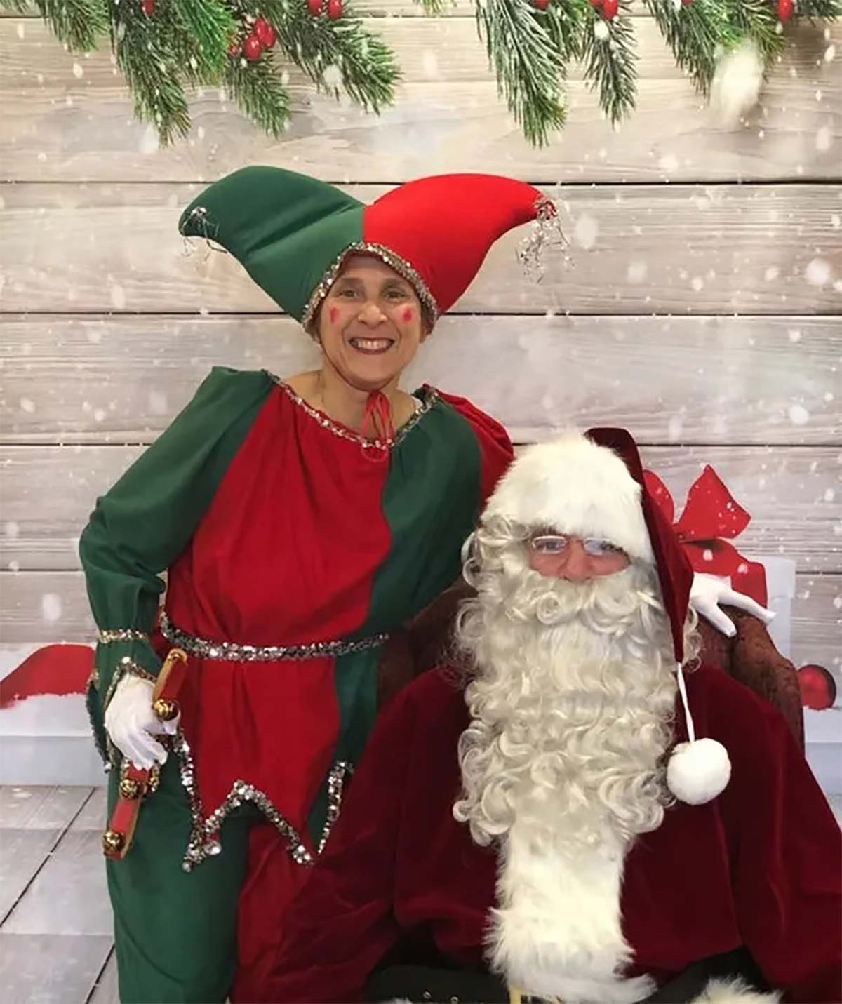Santa Claus with an elf in front of a snowy wooden backdrop, both smiling, festive attire.