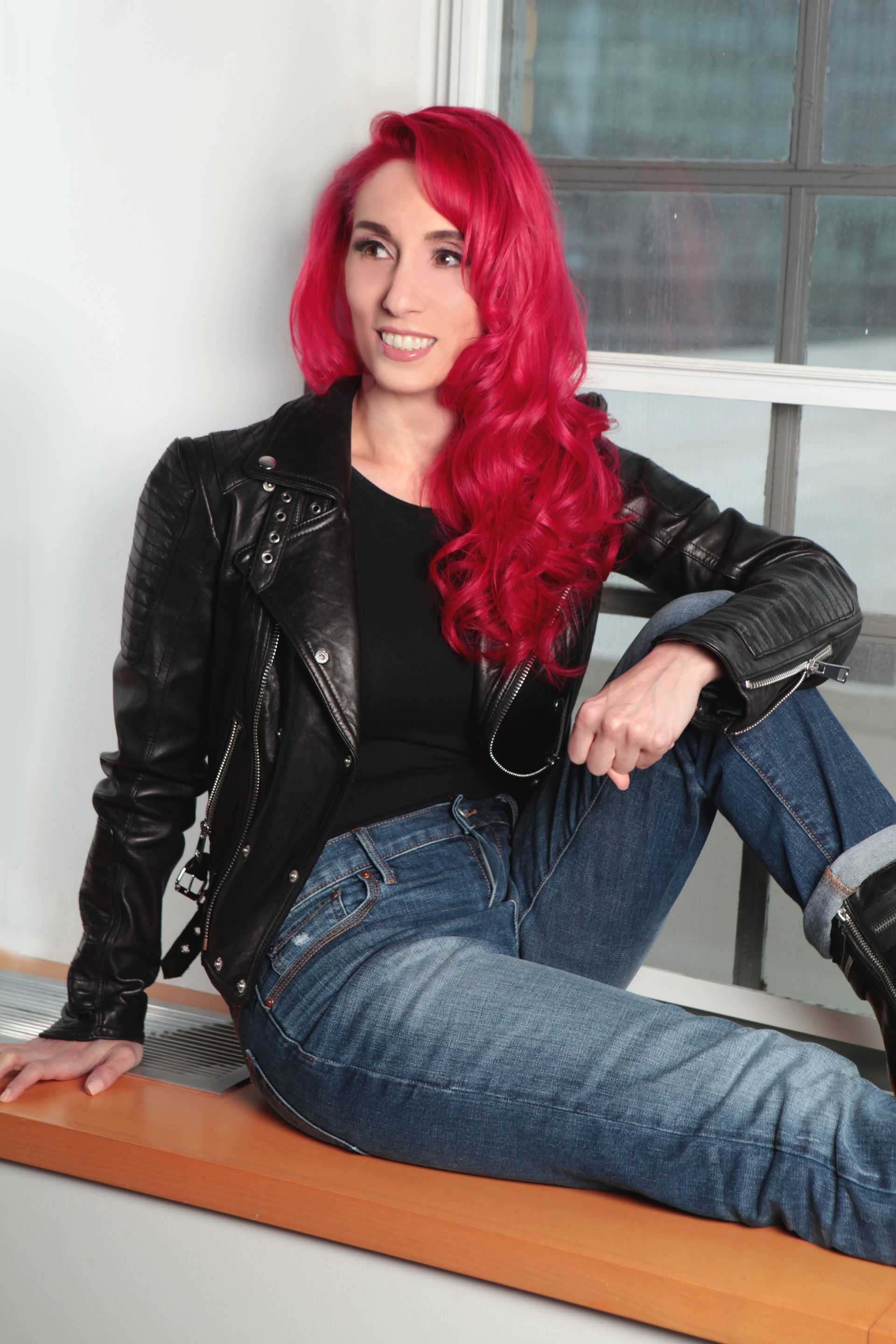 A woman with red hair is sitting on a window sill wearing a black leather jacket and jeans.
