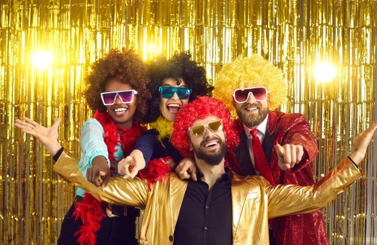 Four people in colorful wigs and sunglasses posing with arms out in front of a gold tinsel backdrop