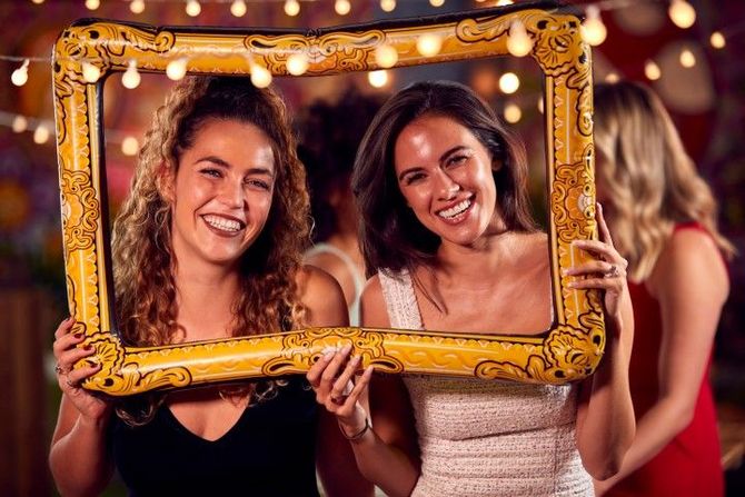 Two women smiling and posing behind a gold decorative frame at a festive event with string lights