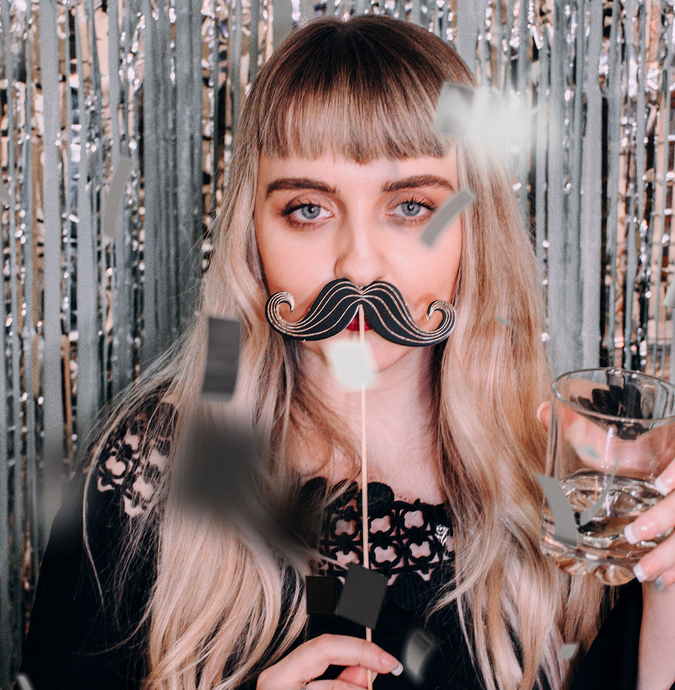 Woman with long blonde hair and a fake mustache, holding a wine glass in front of a silver tinsel backdrop
