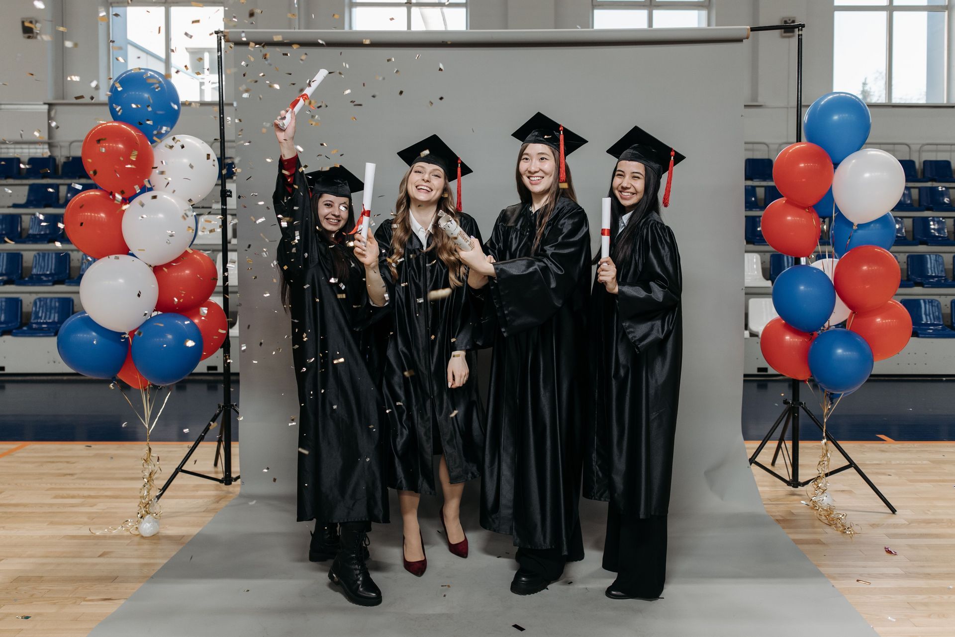 Four graduates in black gowns and caps celebrate in front of balloon columns and a gray backdrop.
