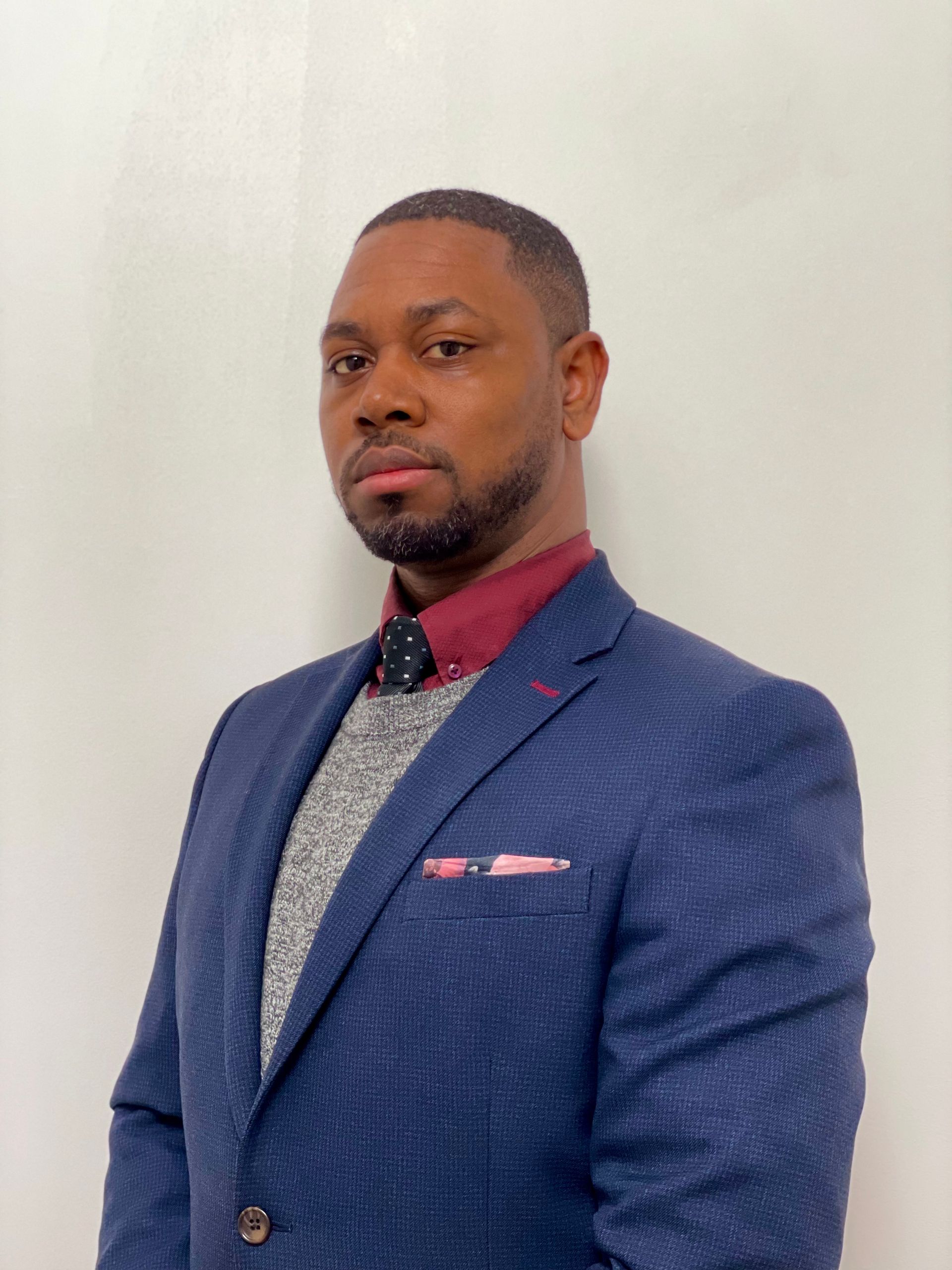 Man in a navy blazer, burgundy shirt, and gray sweater, looking directly at the camera.