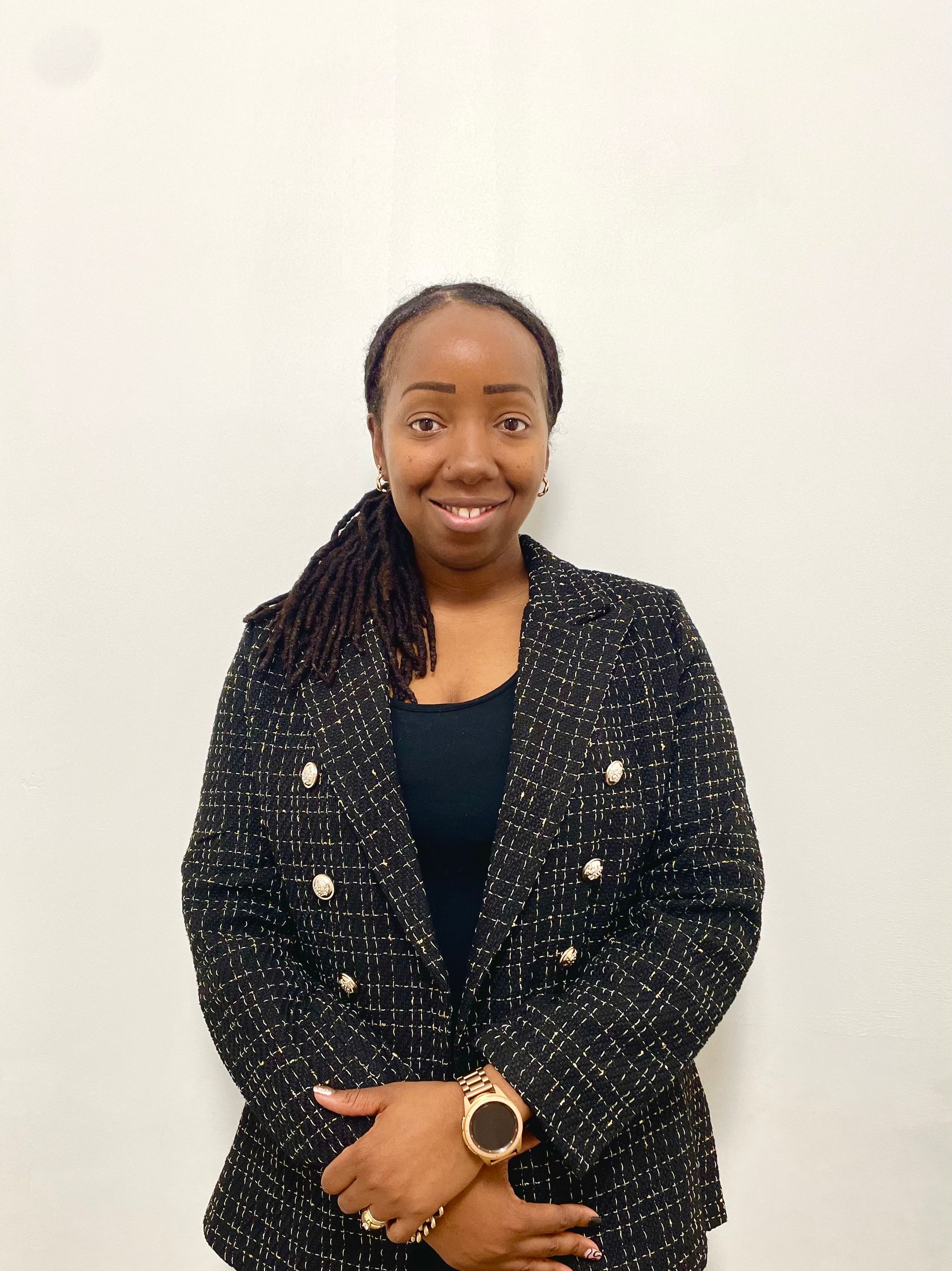 Woman in a black blazer smiling, posing in front of a white wall.