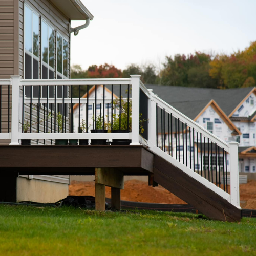 Brown deck with white railing, black spindles, and stairs leading to a yard with houses in the background.