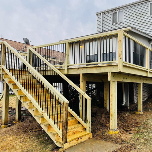 Wooden deck with stairs, black railings, and supporting posts, against a house and overcast sky.