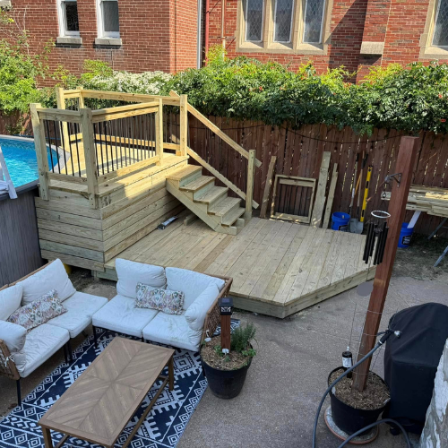 Backyard deck with seating area, stairs, and railing. Wooden construction, surrounded by greenery.