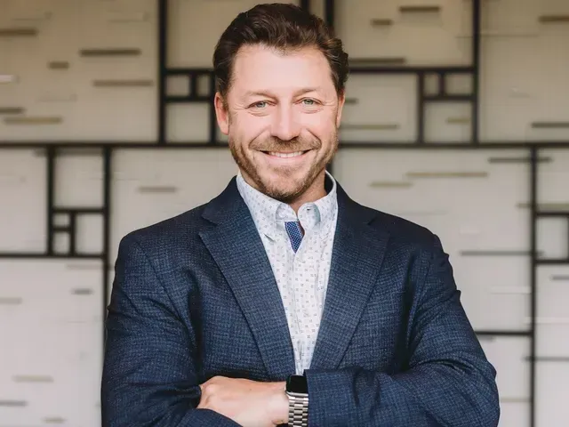 Smiling man in a blue blazer with arms crossed, standing before a geometric wall background
