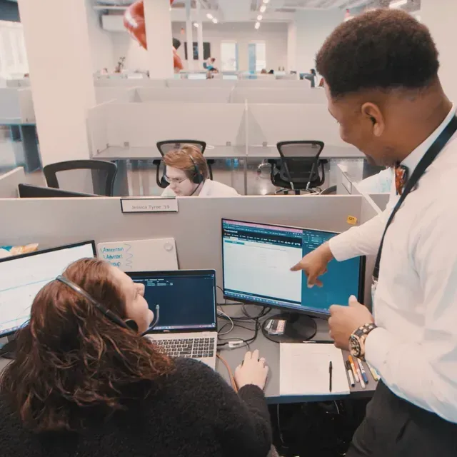 Colleagues reviewing data on office monitors, with one pointing at a screen during a meeting