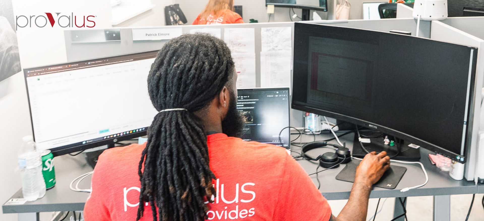 Person seated at dual monitors in an office, wearing a red shirt and working at a desk