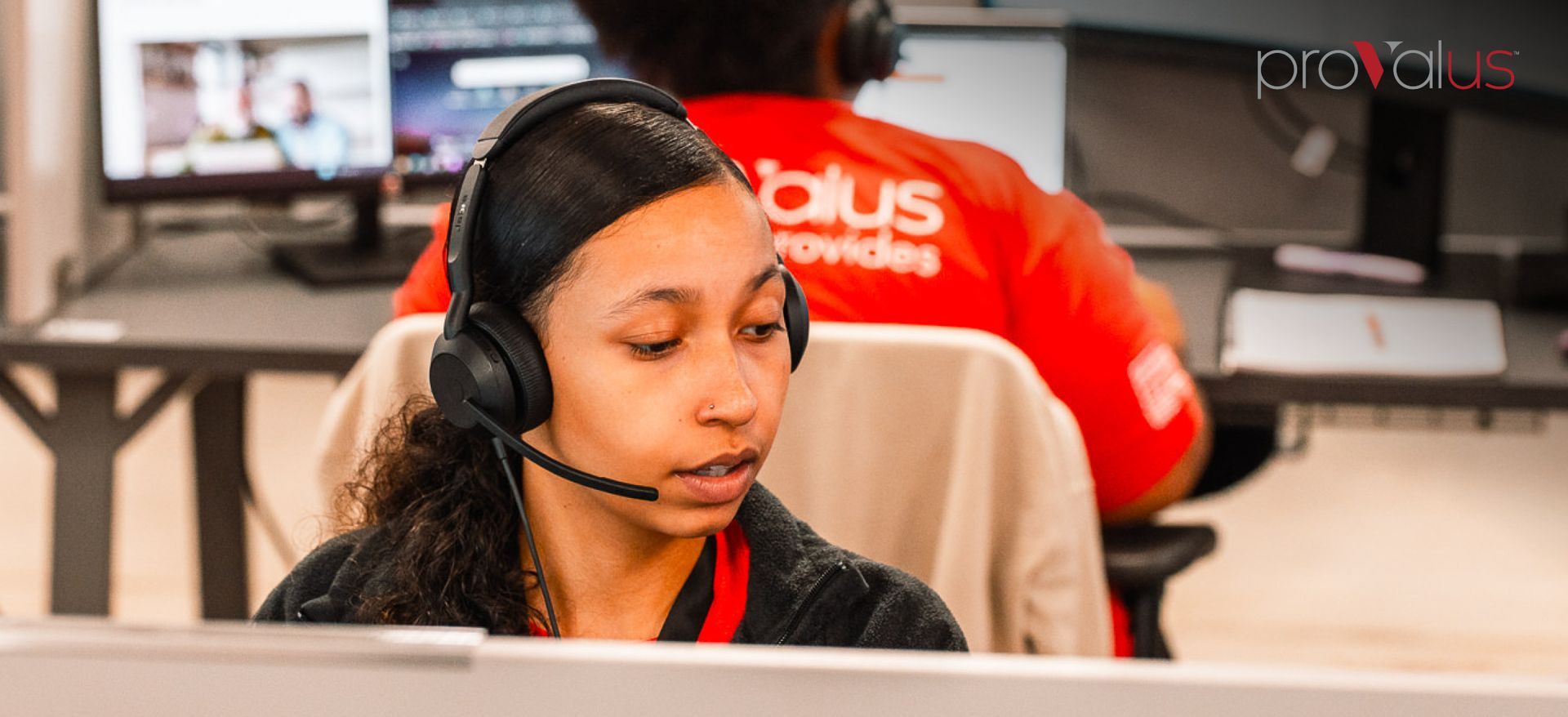 Call center agent wearing a headset at a computer, with a coworker in the background