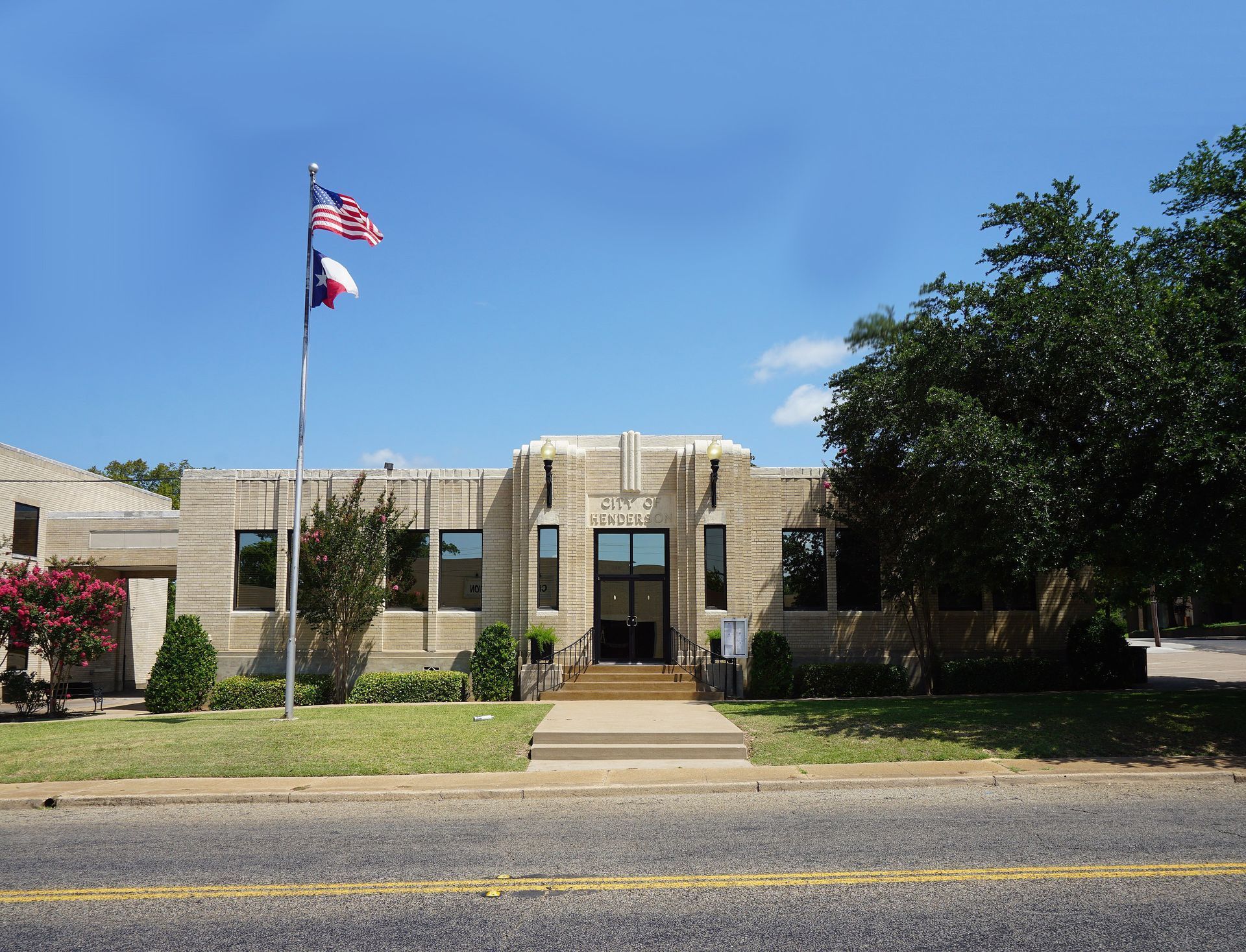 Stone civic building with U.S. and Texas flags, centered walkway, and trees under a clear blue sky