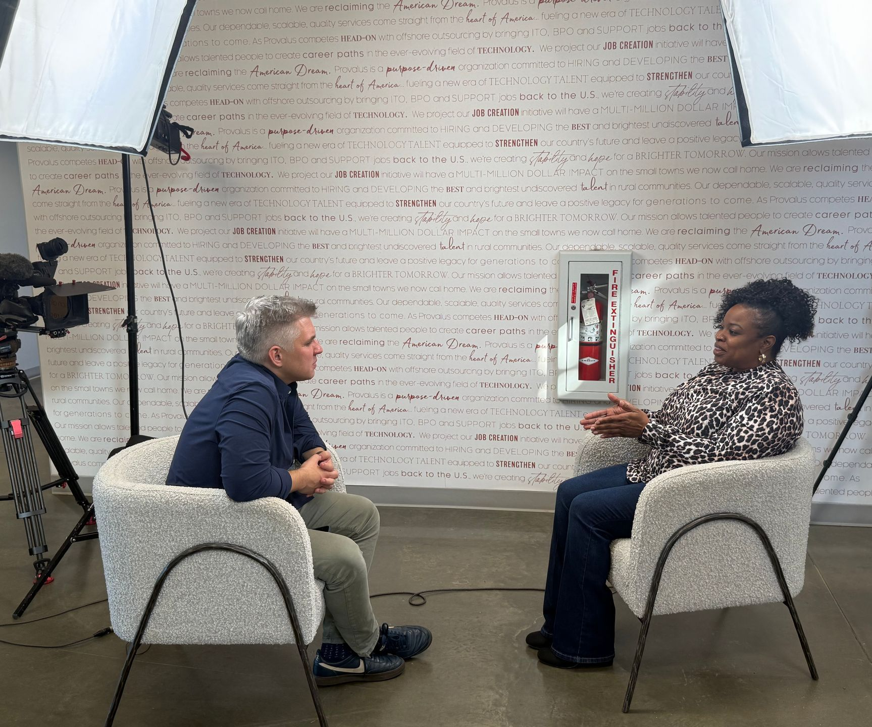Two people seated in a studio interview setup, facing each other with cameras and lighting in view.