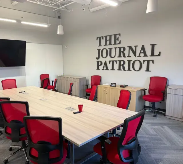 Conference room with red chairs, light wood table, and “THE JOURNAL PATRIOT” wall sign