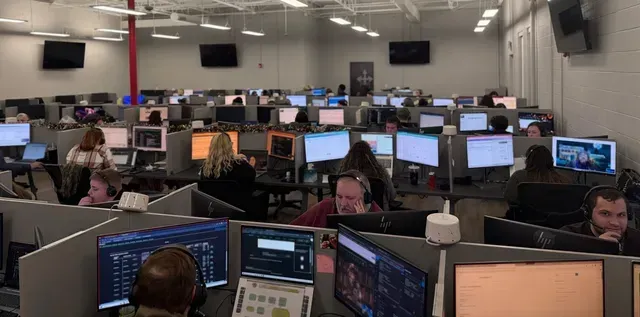 Busy call center with many employees at computers in a large office, lit by overhead lights.