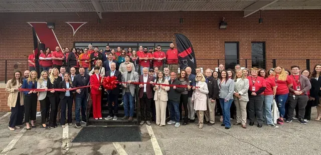 Group of people in red and black standing outside a brick building, holding a ribbon for a ceremony