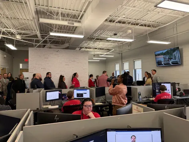 People waiting in a busy office with cubicles, computers, and a wall-mounted TV.