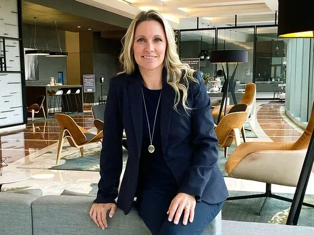 Woman sitting in a modern lounge, wearing a navy blazer and jeans, with floor-to-ceiling windows behind her