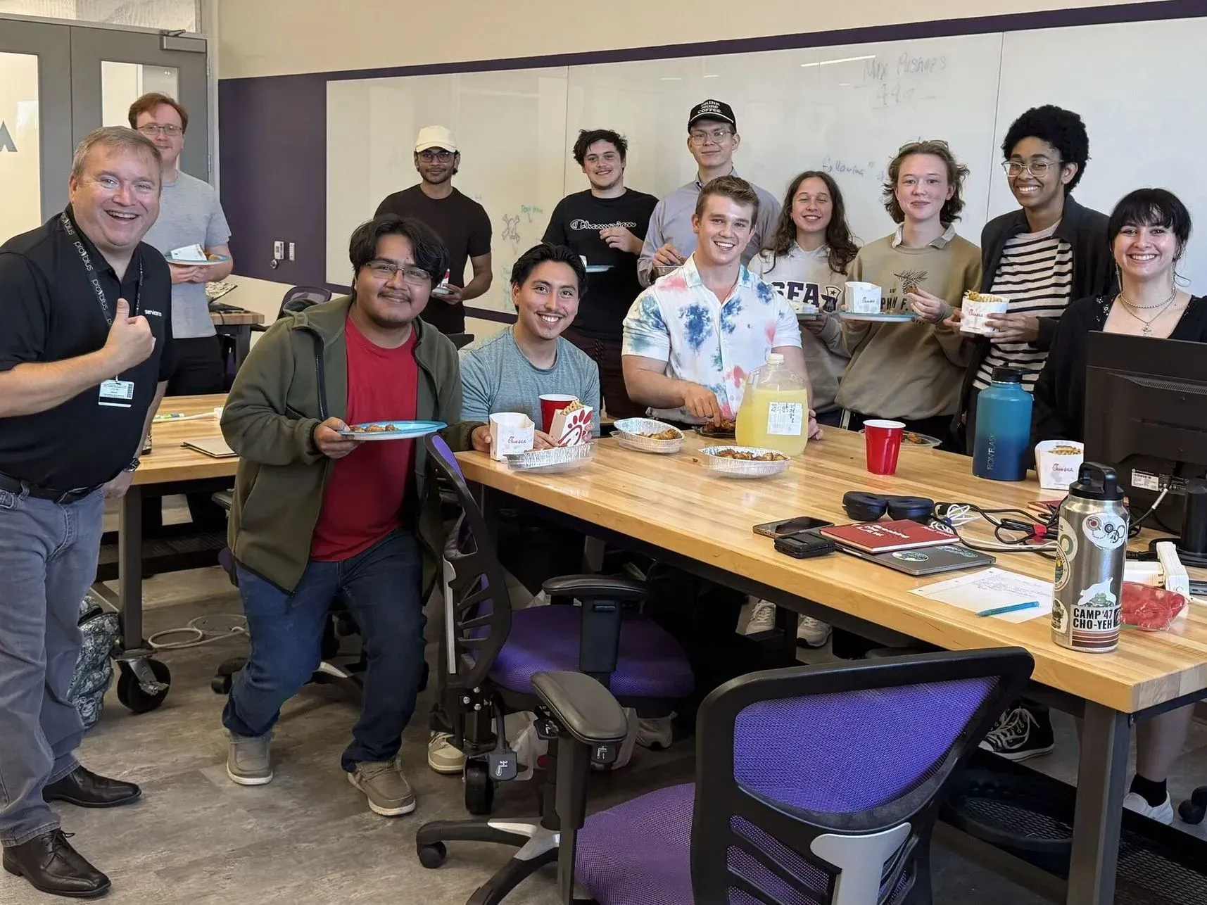Group of people smiling in a classroom around a wooden table, holding plates and cups during a celebration