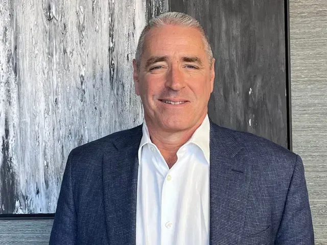 Smiling man in a blue blazer and white shirt standing before a gray textured wall