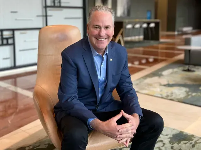 Smiling man in a blue suit seated in a tan chair in a modern office lobby.