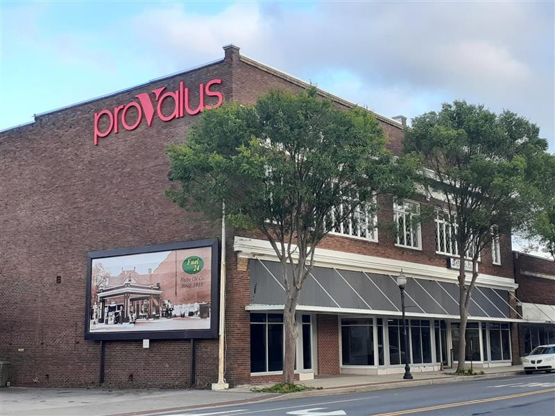 Brick storefront with red “provalus” sign, trees, and parked cars along the street