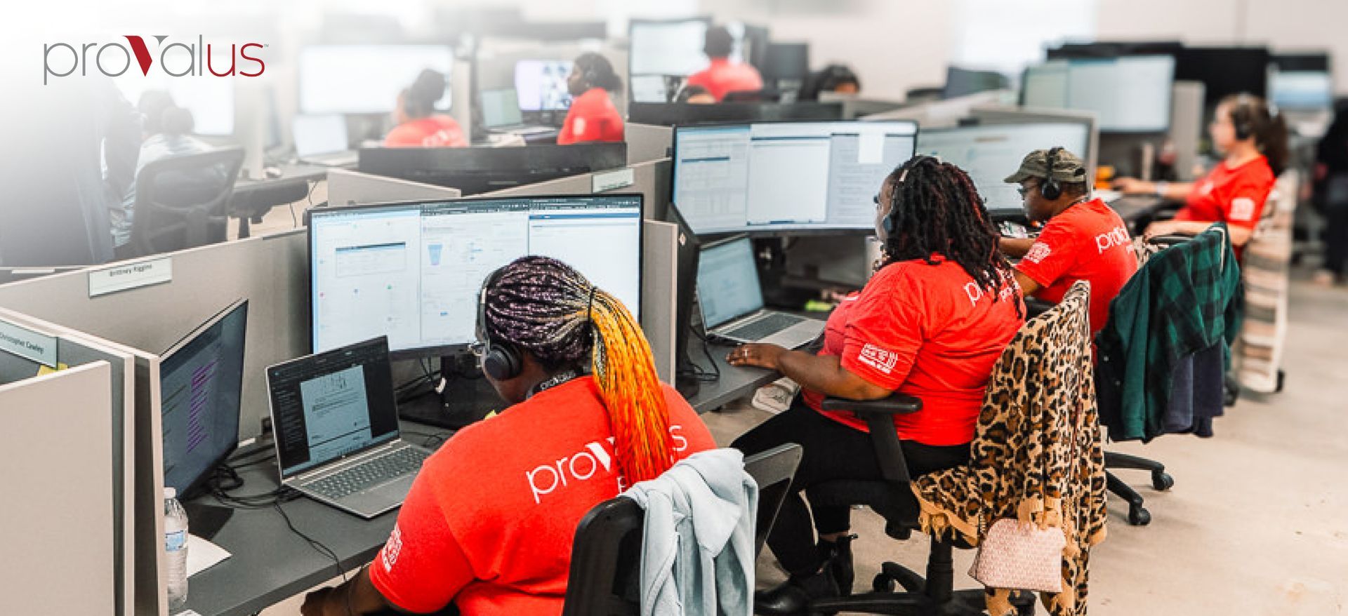 People working at desktop computers in a bright call center with red shirts and rows of monitors