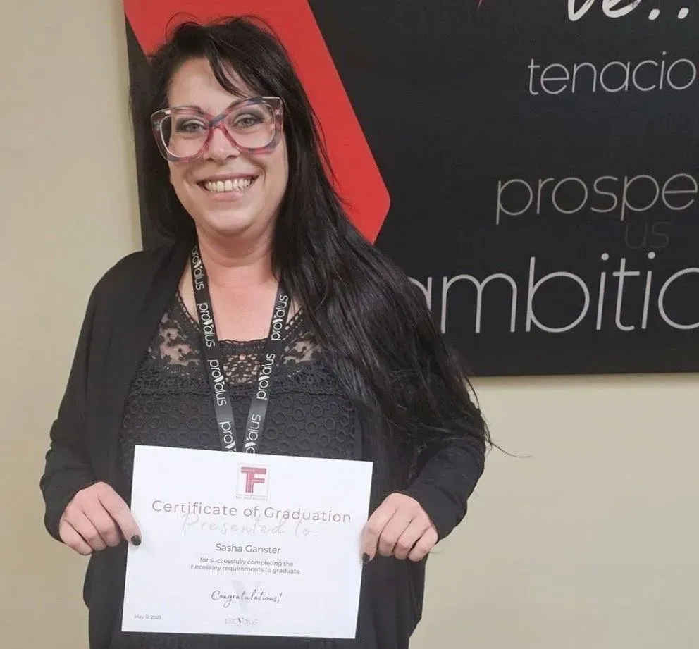 Woman smiling and holding a certificate in front of a wall sign with motivational words.