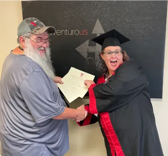Two people shaking hands, one in a graduation cap and gown, holding a certificate in front of an Adventurous sign.