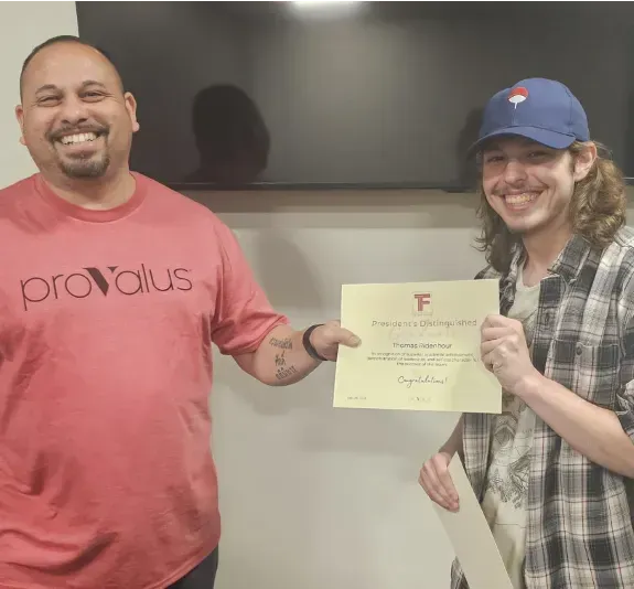 Two smiling people hold a certificate in front of a wall-mounted TV.