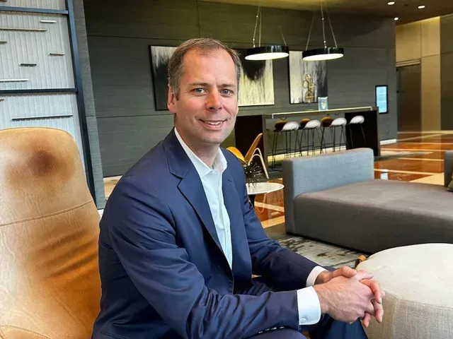 Man in a blue suit seated in a modern lounge with warm lighting and upholstered chairs