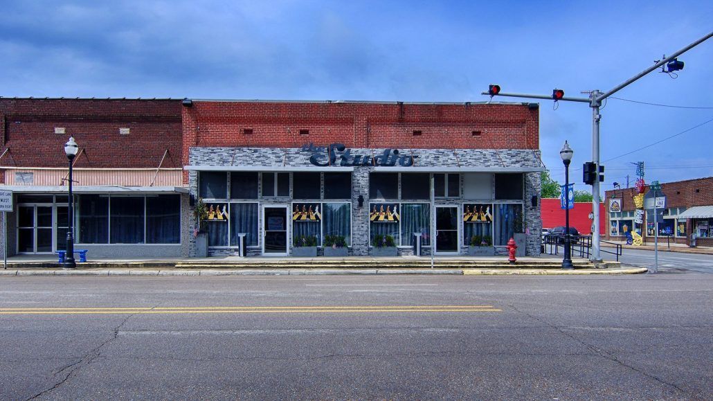 Small storefronts with red roofs and awnings along an empty street under a blue sky.