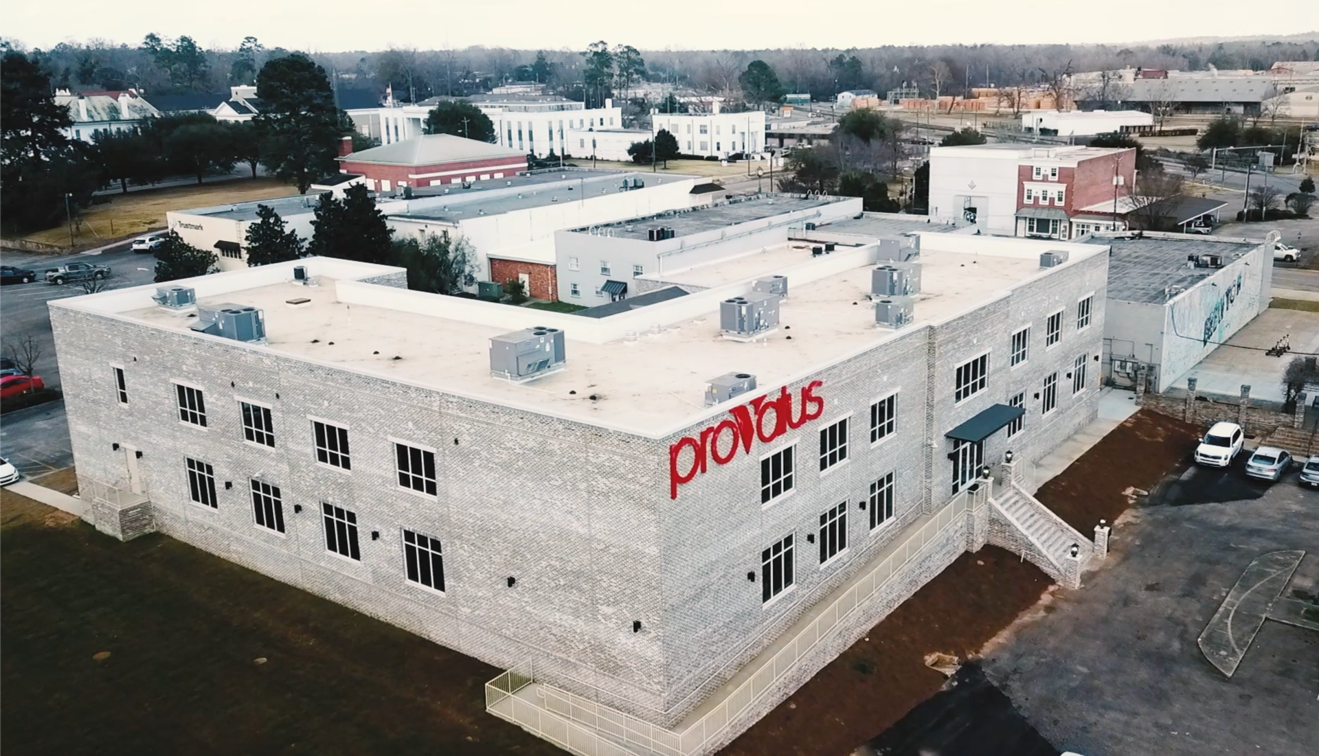 Aerial view of a white industrial building with red sign, surrounded by parking and neighboring buildings.