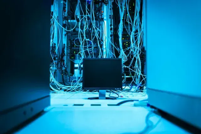 Computer monitor in a blue-lit server room with tangled cables and illuminated equipment behind it