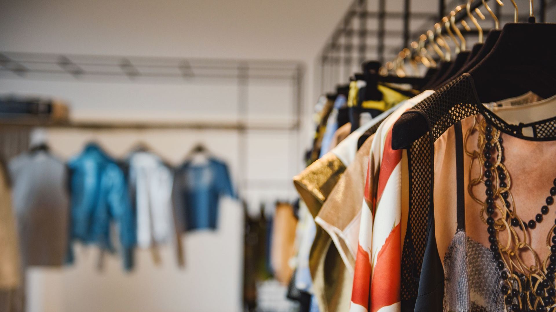 A clothing rack with various garments, including a patterned top with long necklaces in the foreground and blue jackets.