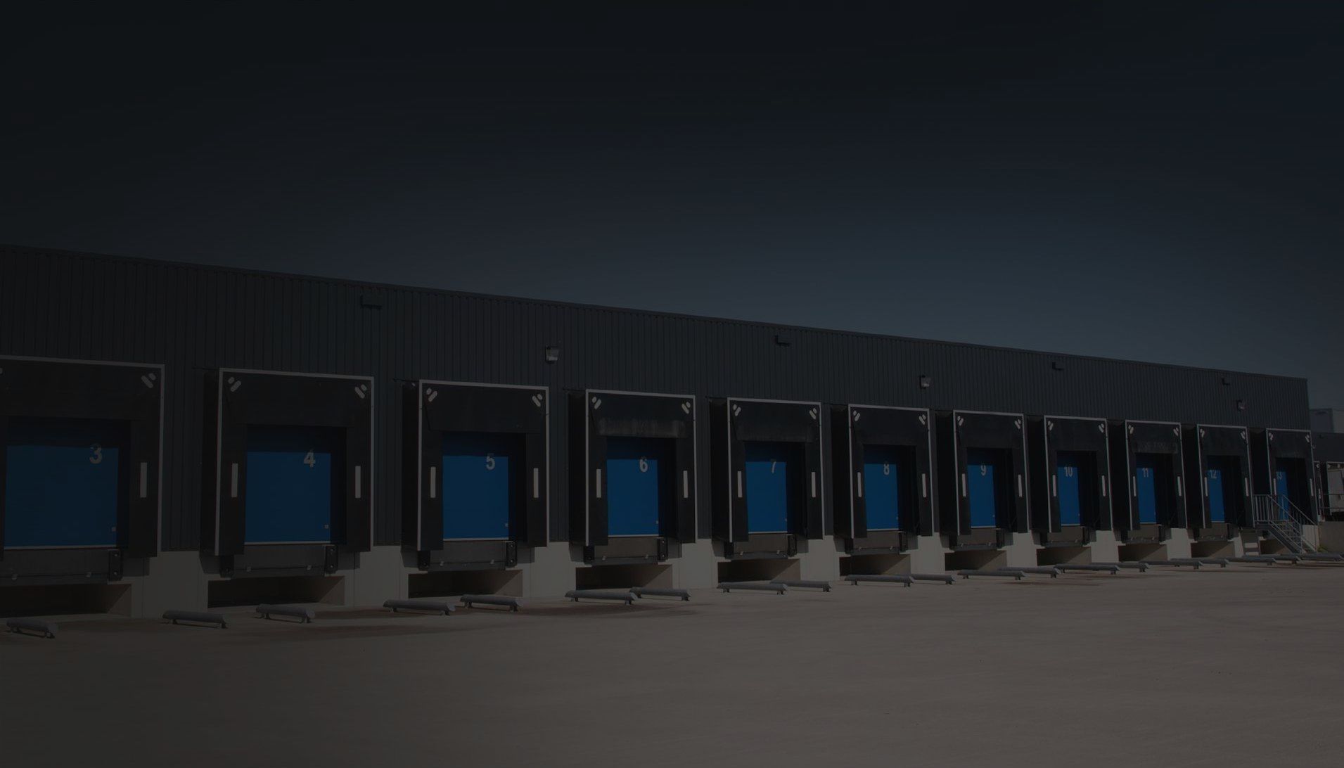 A row of shipping loading docks with blue door seals against a dark metal industrial building at dusk.