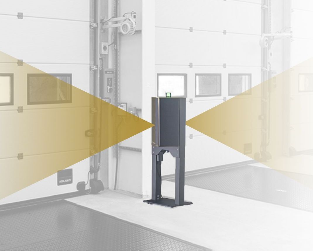 A black bollard sensor stands in a loading bay, projecting two orange light beams horizontally to detect movement.