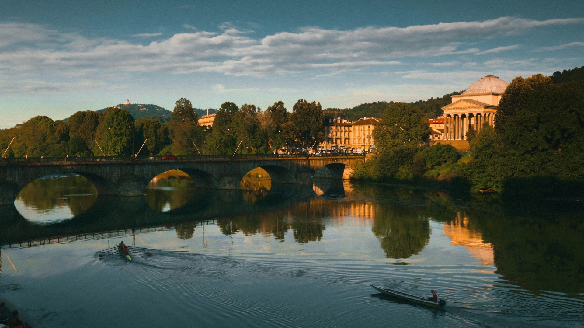 Paesaggio fluviale a Torino, Italia, con una barca, un ponte e degli edifici che si riflettono nell'acqua, sotto un cielo blu e dorato.