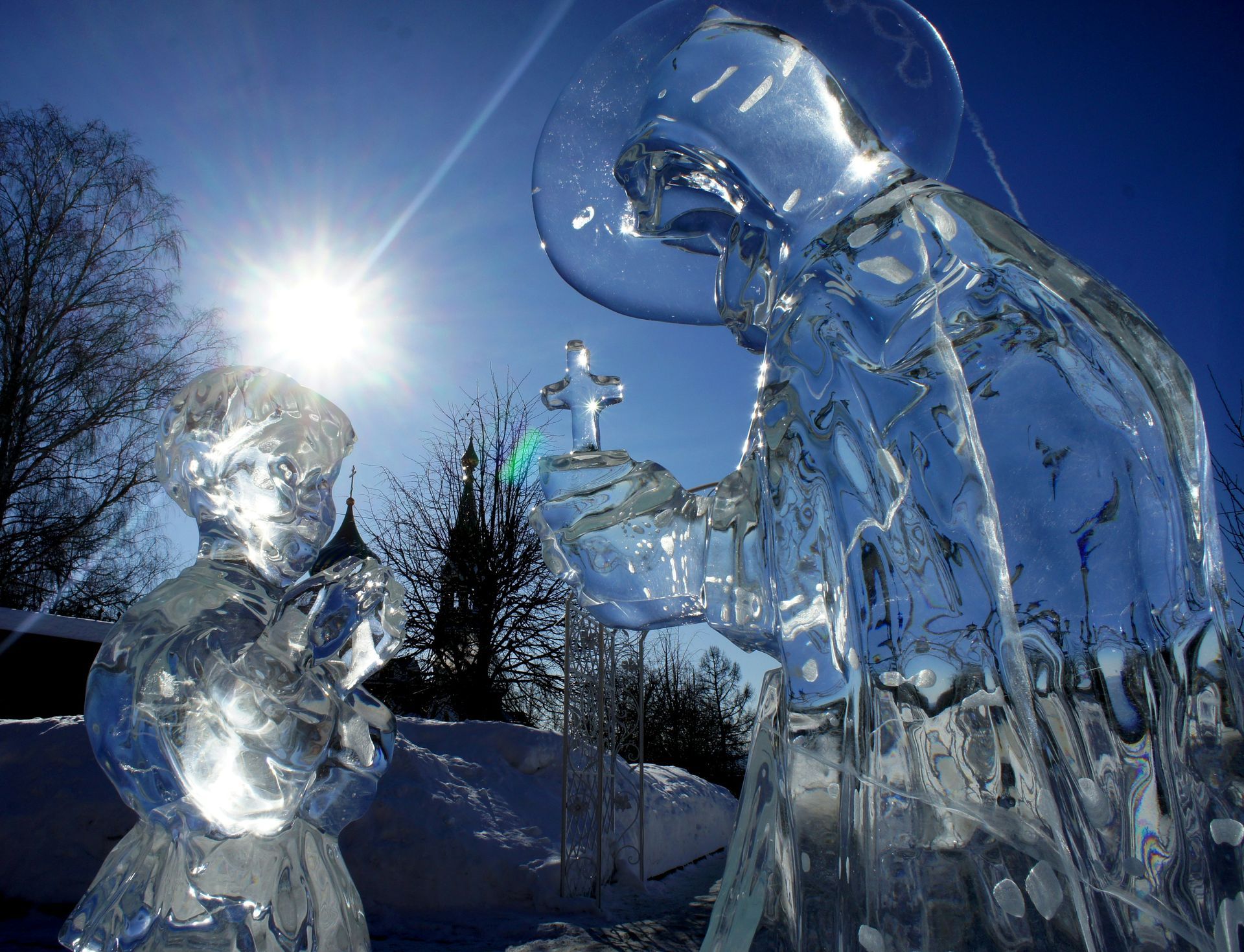 Scultura di ghiaccio raffigurante un santo che porge una croce a un bambino; sullo sfondo splende il sole.