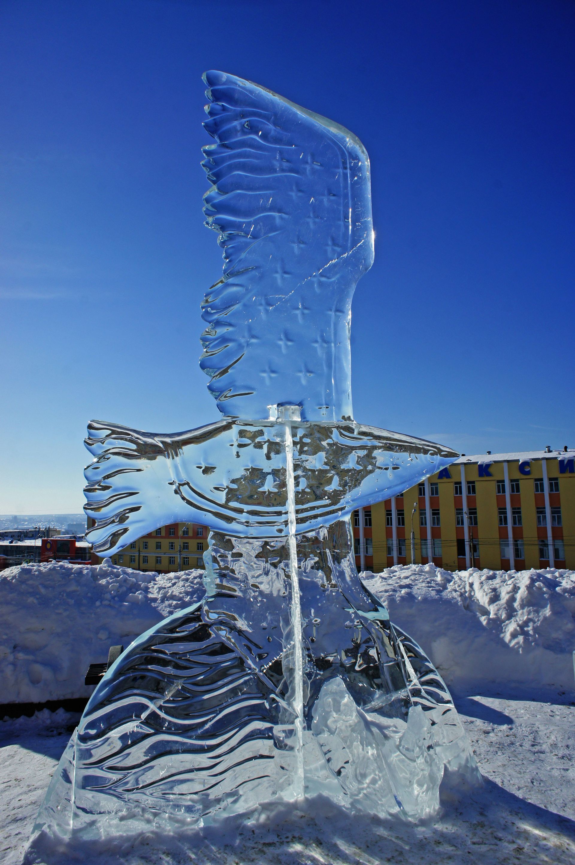 Scultura di ghiaccio raffigurante una figura simile a un uccello con le ali, sulla neve, con un cielo azzurro e degli edifici sullo sfondo.