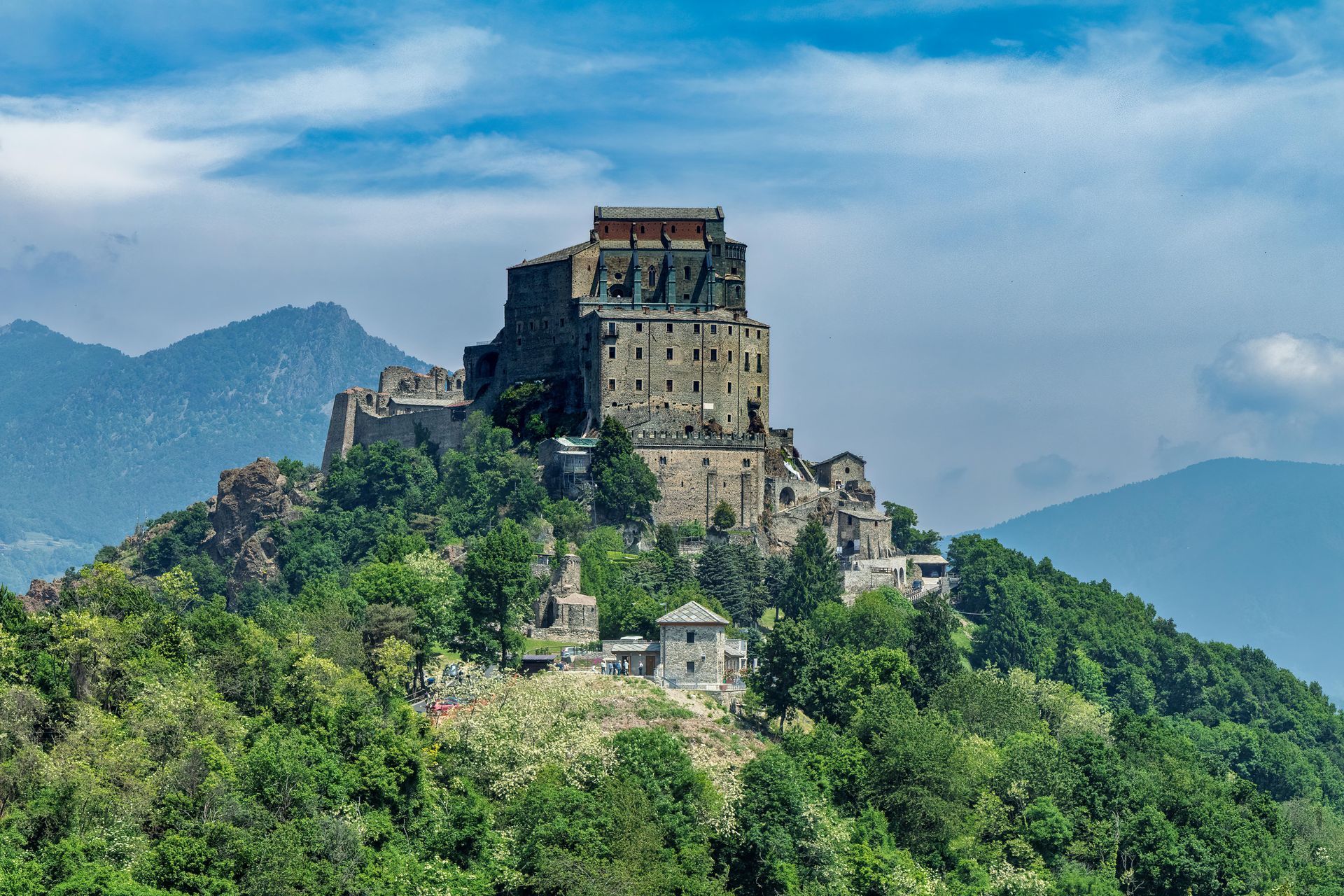Monastero medievale in cima a una collina, circondato da alberi e montagne, sotto un cielo azzurro.