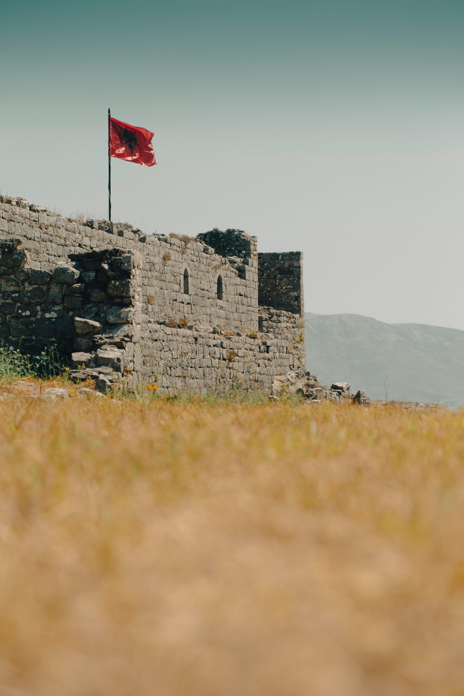 Castello in pietra in rovina con la bandiera albanese che sventola sopra, che domina un paesaggio arido ed erboso.