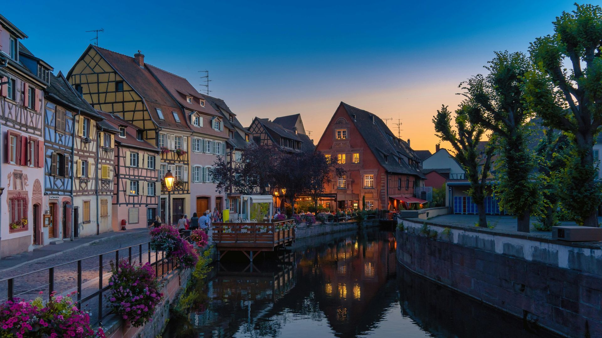 Canale fiancheggiato da edifici colorati a Colmar, in Francia, che riflette le luci della sera; fiori in primo piano.