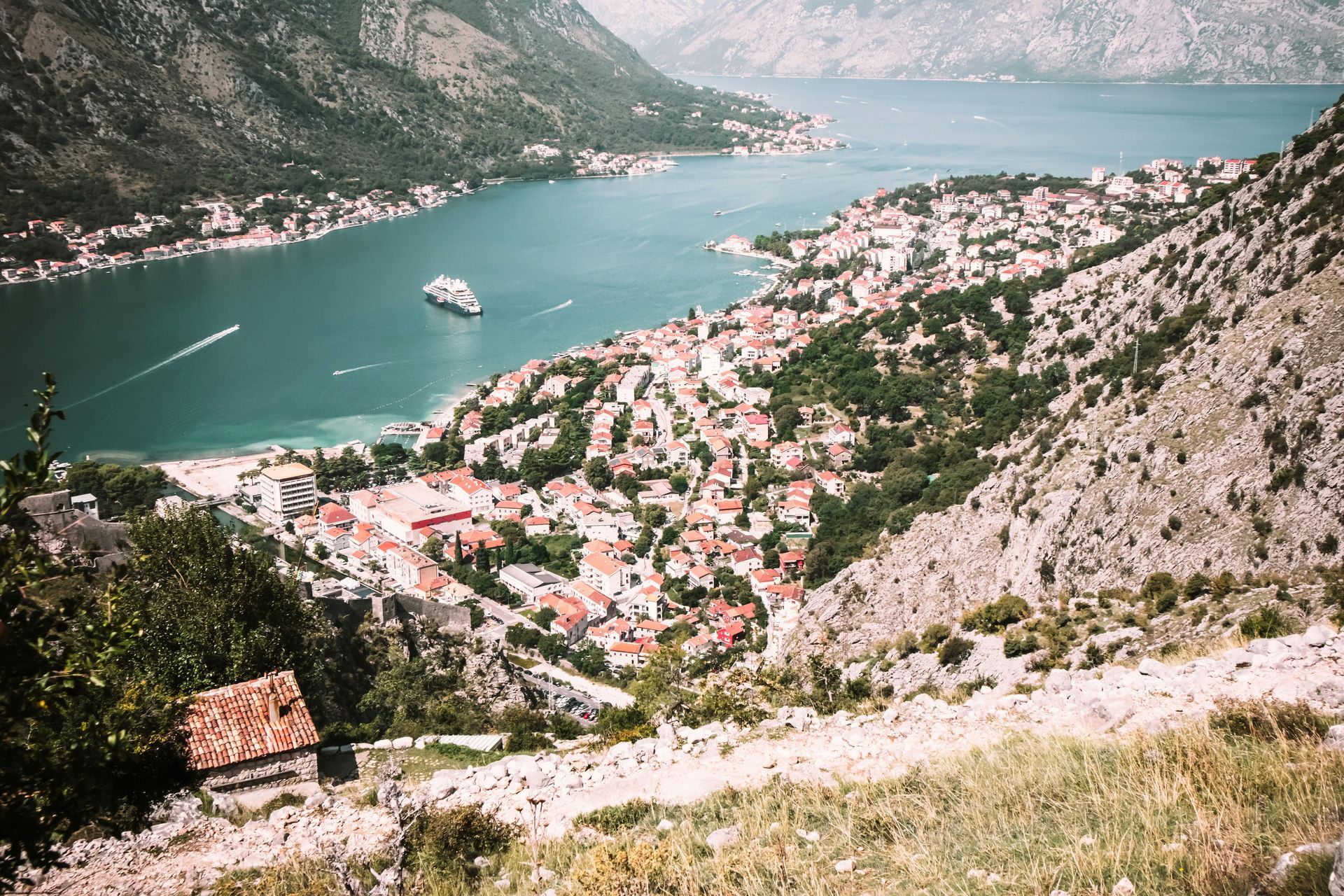 Città costiera immersa in acque turchesi, con le montagne sullo sfondo. Edifici dai tetti rossi, barche sull'acqua.