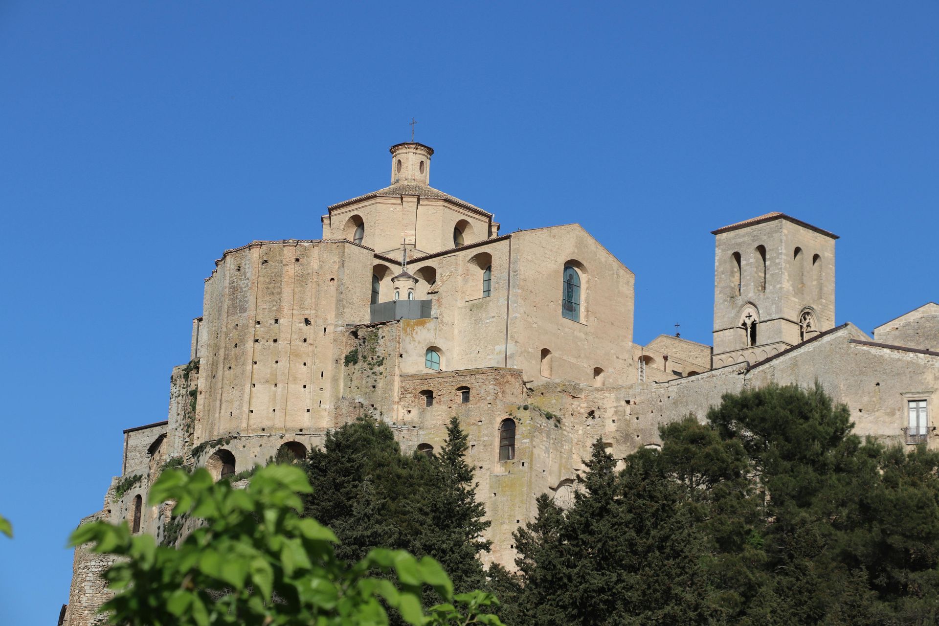 Struttura di un castello in pietra contro un cielo azzurro, arroccato su una collina con alberi in primo piano.