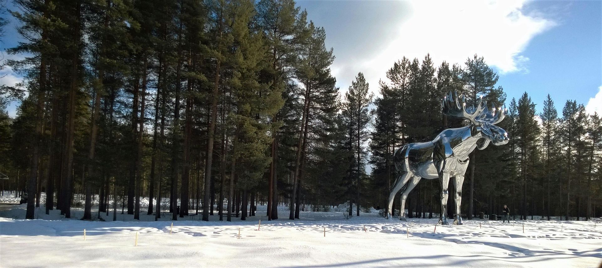 Una scultura in metallo raffigurante un alce in una foresta innevata. Sullo sfondo si vedono gli alberi, sopra il cielo azzurro con le nuvole.